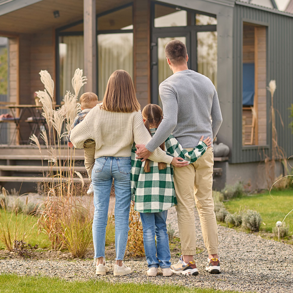 Happy family. View from back of man woman with child and girl standing together hugging and admiring their cozy home on fine autumn day